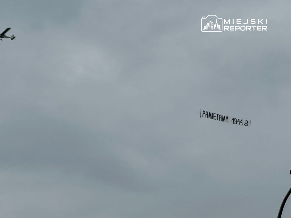 Samolot ciągnie za sobą baner z napisem "PAMIĘTAMY 1944" na szarym niebie.
