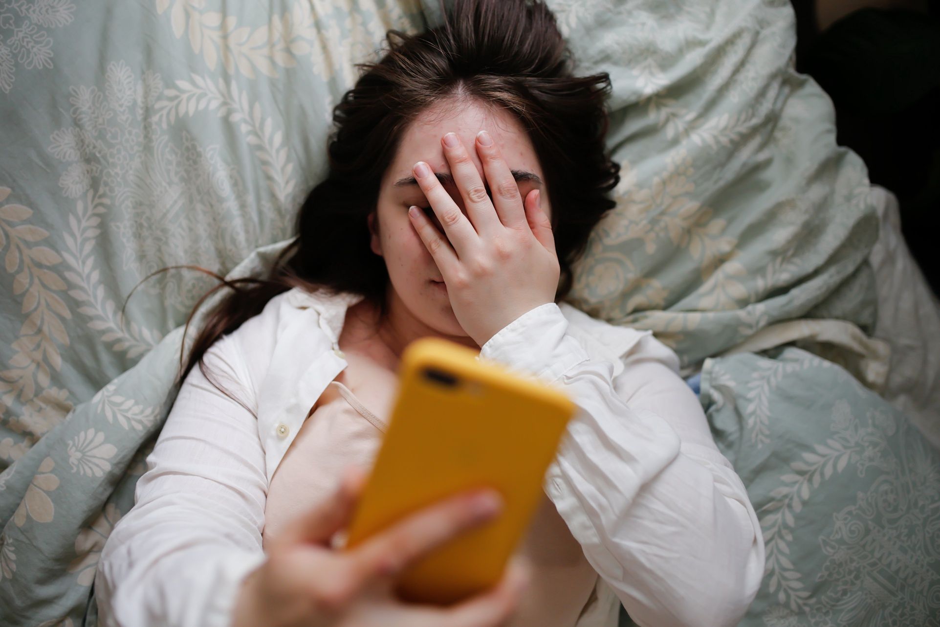 Sleepy overweight young woman with flowing hair using phone on bed, close face palm. Top viewPlus size young woman in home bedroom on bed with phone. Woman covering her eyes with her handNatalia  Lebedinskaia