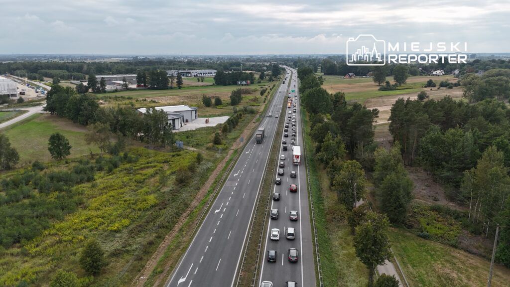 Ruch samochodowy na szerokiej drodze, z długim korkiem na jednym pasie, w otoczeniu zieleni i zabudowy przemysłowej.