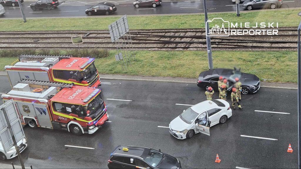 Strażacy w kaskach rozmawiają obok uszkodzonego białego samochodu na ruchliwej drodze, obok dwóch wozów strażackich.