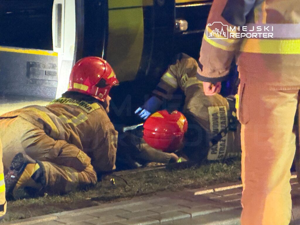 Dwóch strażaków w hełmach, leżących na trawie, udziela pomocy osobie w pobliżu pojazdu.