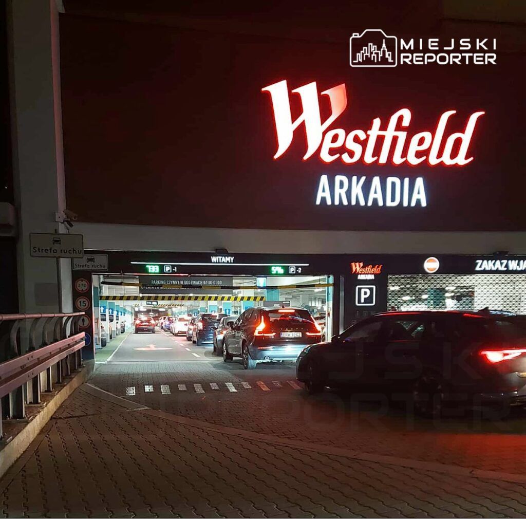 Ruchliwy wjazd do parkingu podziemnego z widocznym znakiem „Westfield Arkadia” oraz samochodami czekającymi na wjazd.