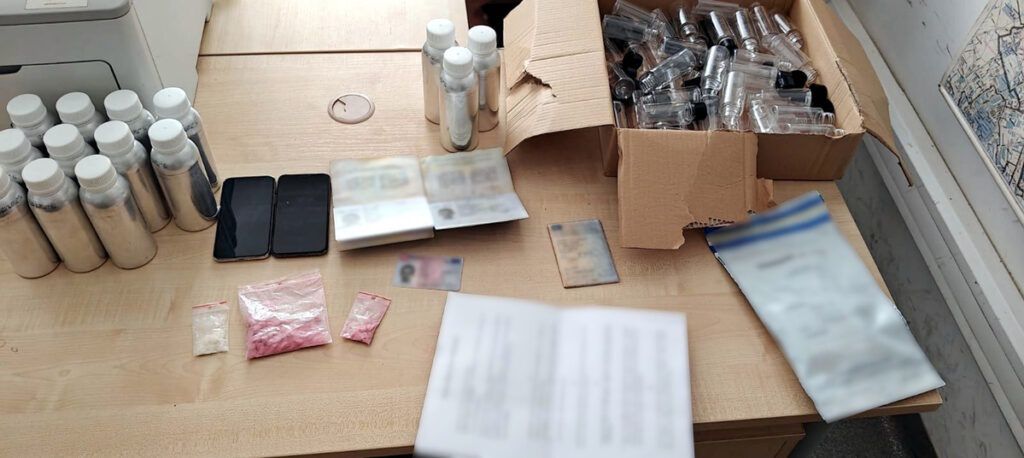 Na blacie biurka znajdują się butelki, dwa telefony, pakunki z substancjami oraz dokumenty tożsamości.