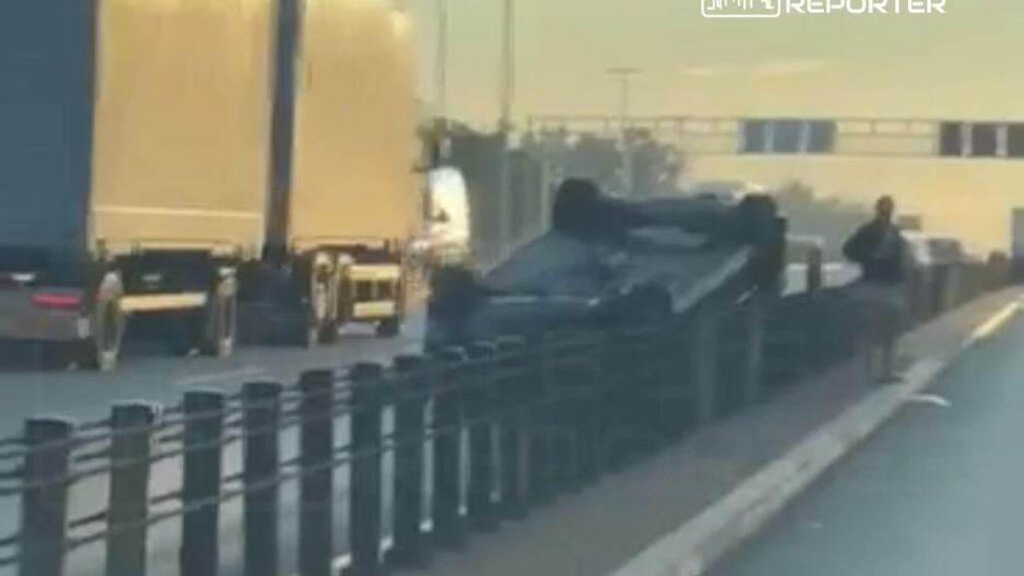 Na drodze leży przewrócone auto, obok stoi mężczyzna, a w tle przejeżdżają ciężarówki.