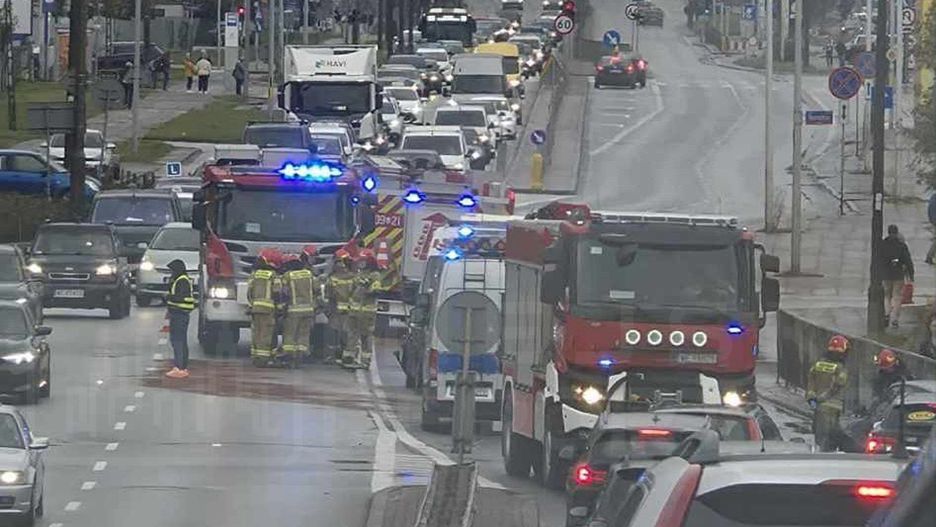 Grupa strażaków w mundurach omawia sytuację obok wozu strażackiego na ruchliwej ulicy, w tle zablokowany ruch samochodowy.