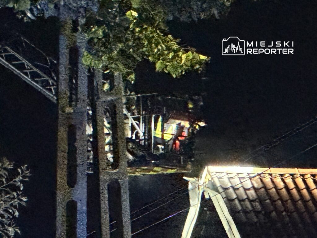 Wysoka drabina strażacka z włączonymi światłami, z której widać sylwetki strażaków, w nocy nad dachem budynku.