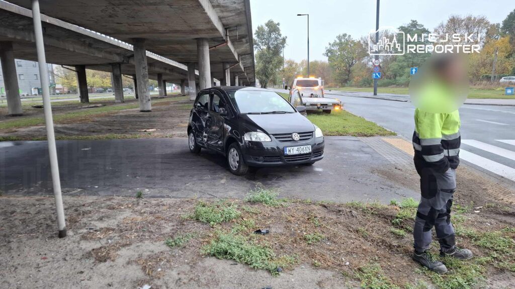 Czarny samochód osobowy z uszkodzeniami stoi pod wiaduktem, obok pracownik w odzieży roboczej obserwuje sytuację.