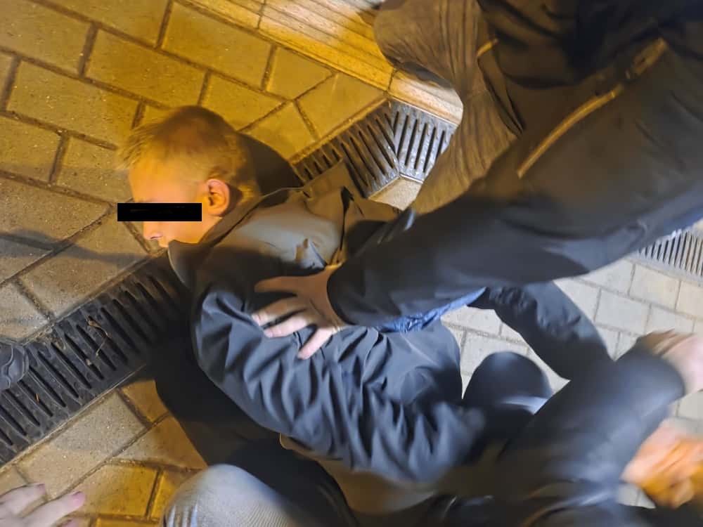 Mężczyzna w ciemnej odzieży leży na chodniku, podczas gdy dwie osoby go przytrzymują.
