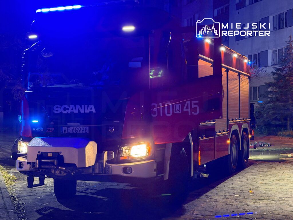 Czerwony wóz strażacki z włączonymi sygnałami świetlnymi stoi na oświetlonym chodniku w pobliżu budynku mieszkalnego.