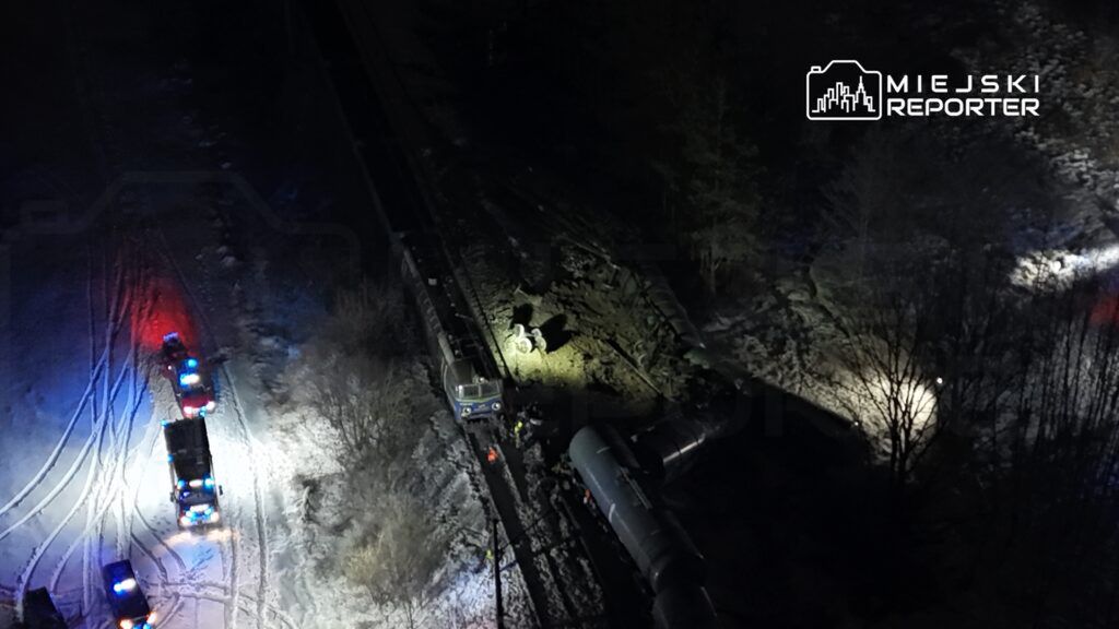 W nocy, zespół ratunkowy pracuje przy przewróconym wagonie kolejowym, otoczony przez pojazdy z włączonymi sygnałami świetlnymi.