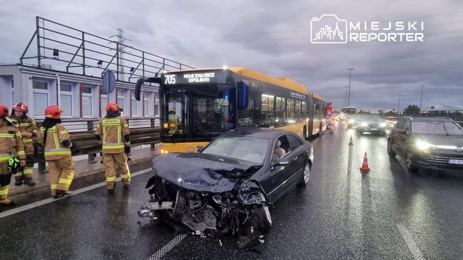 Grupa strażaków w kombinezonach ratunkowych sprawdza uszkodzony samochód osobowy na jezdni obok autobusu komunikacji miejskiej.