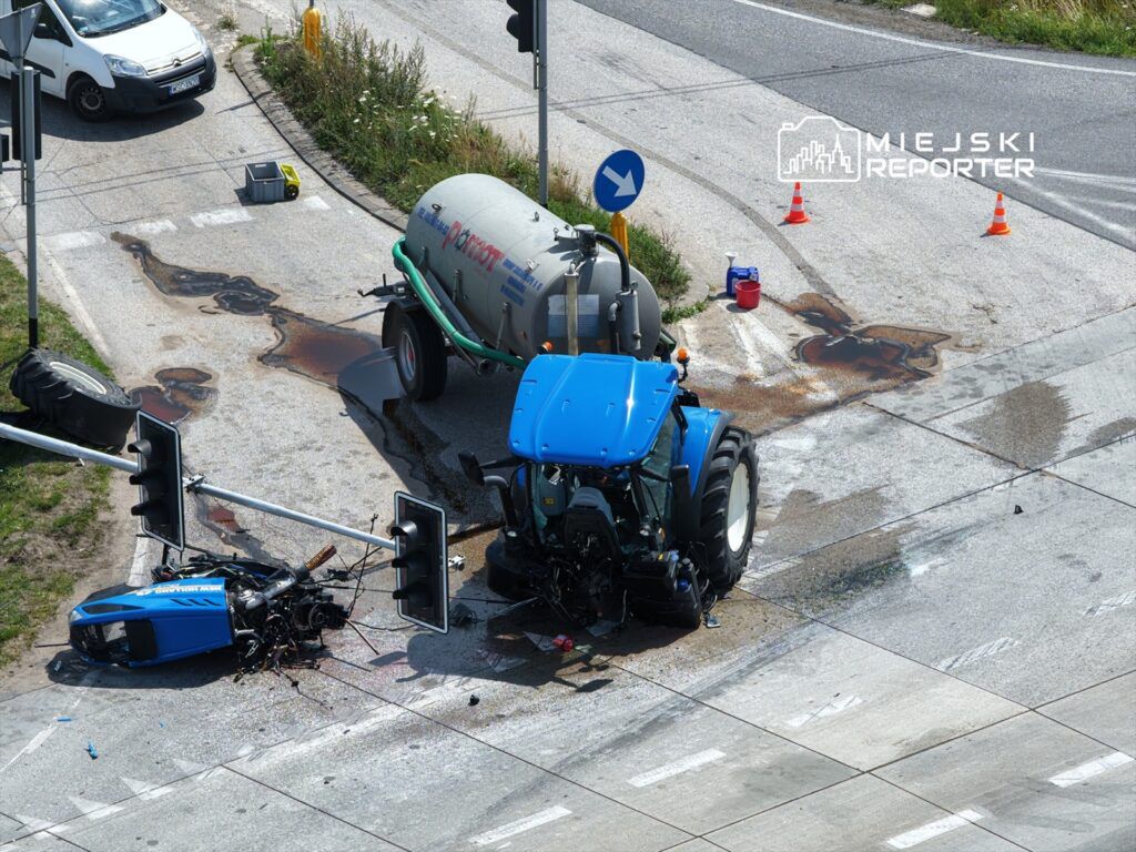 Traktor z przewróconą kabiną leży na drodze, obok rozlane płyny, a w tle widać znaki drogowe i pojazdy.