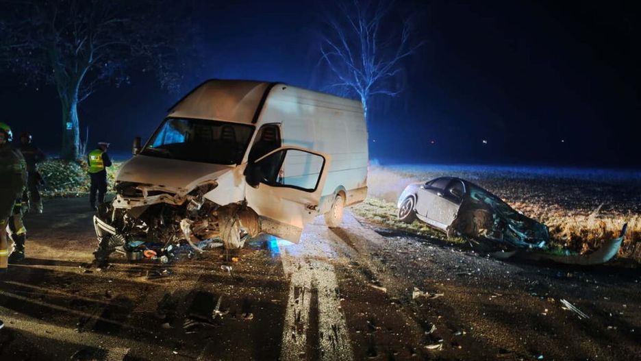 W nocy dwa rozbite pojazdy, dostawczy i osobowy, leżą na drodze, a obok pracują strażacy i policjanci.
