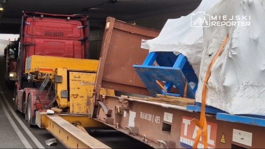 Czerwony ciężarowy pojazd z załadunkiem przejeżdża przez tunel, obok widoczny jest transport kontenerów.