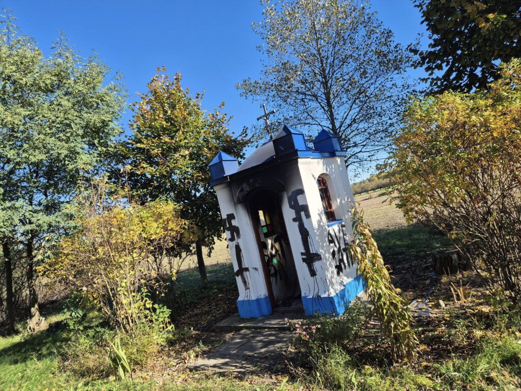 Kapliczka z niebieskim dachem otoczona kolorowymi krzewami w słoneczny dzień na tle jasnego nieba.