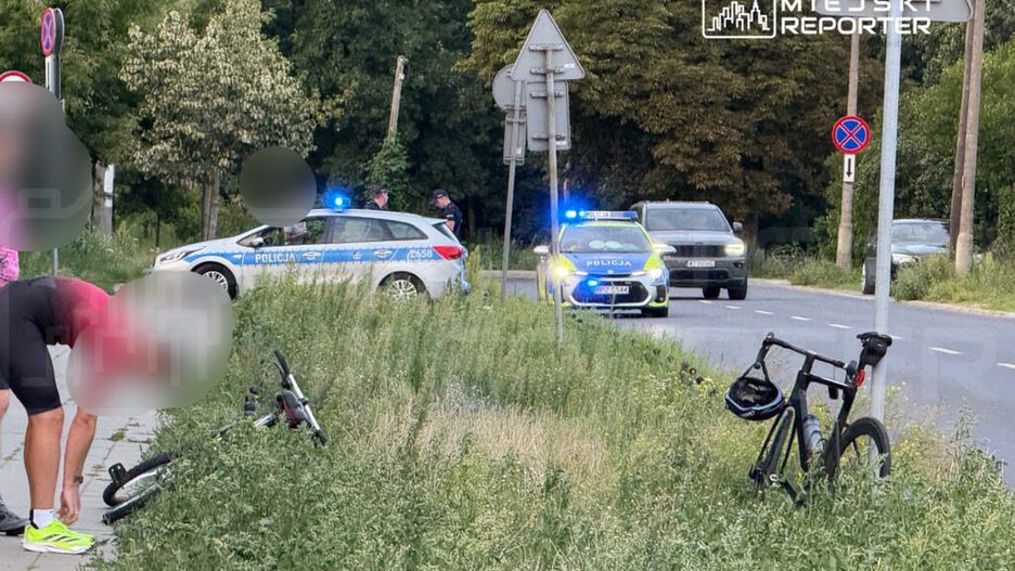 Mężczyzna w różowej koszulce schyla się przy rowerze na poboczu drogi, obok policyjny radiowóz z włączonymi sygnałami.