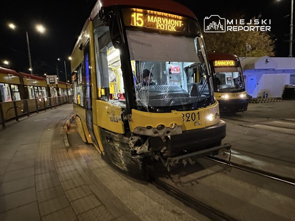 Złamany tramwaj z uszkodzonym przodem stoi na przystanku w nocy, obok inny tramwaj w tle.