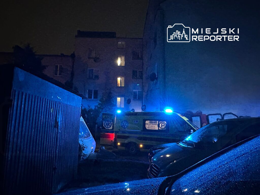 Nocna scena z niebieskimi światłami karetki w wąskiej uliczce, otoczonej budynkami i zaparkowanymi samochodami.