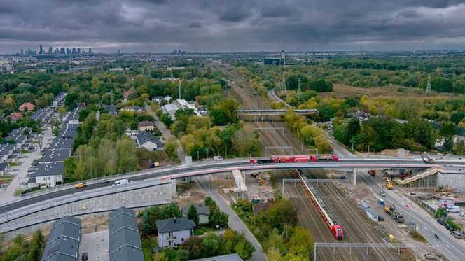 Zabudowa mieszkaniowa sąsiadująca z nowym wiaduktem kolejowym, nad torami, w otoczeniu zieleni i miejskiego krajobrazu.