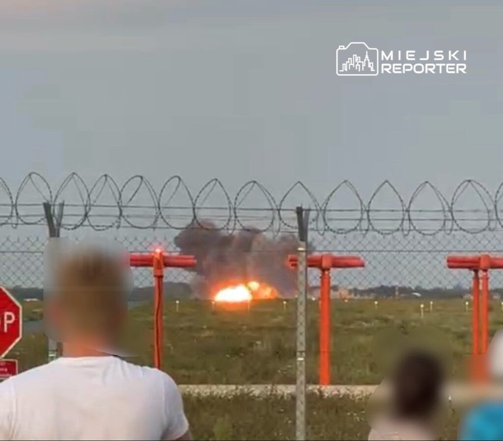 Osoby stojące za ogrodzeniem z drutu kolczastego obserwują wybuch na terenie lotniska.