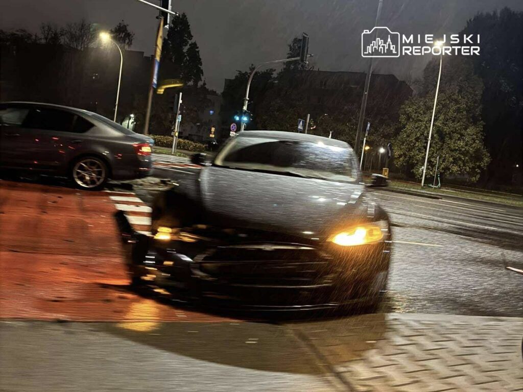 Czarny samochód osobowy z uszkodzoną przednią częścią stoi na skrzyżowaniu w deszczową noc.
