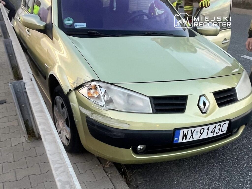 Zielony samochód marki Renault uszkodzony na skraju jezdni, obok stoją strażacy.