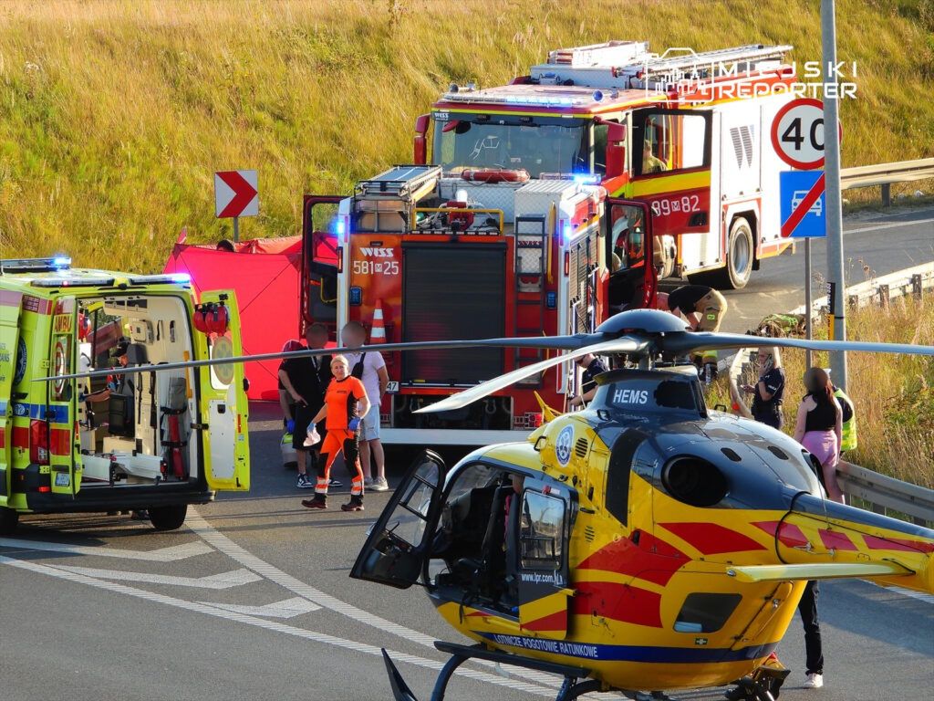 Na drodze, w pobliżu wypadku, zespół ratunkowy pracuje przy ambulansie i śmigłowcu, a strażacy rozstawiają sprzęt.