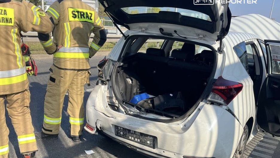 Dwóch strażaków w mundurach obserwuje uszkodzone białe auto z otwartym bagażnikiem na drodze.