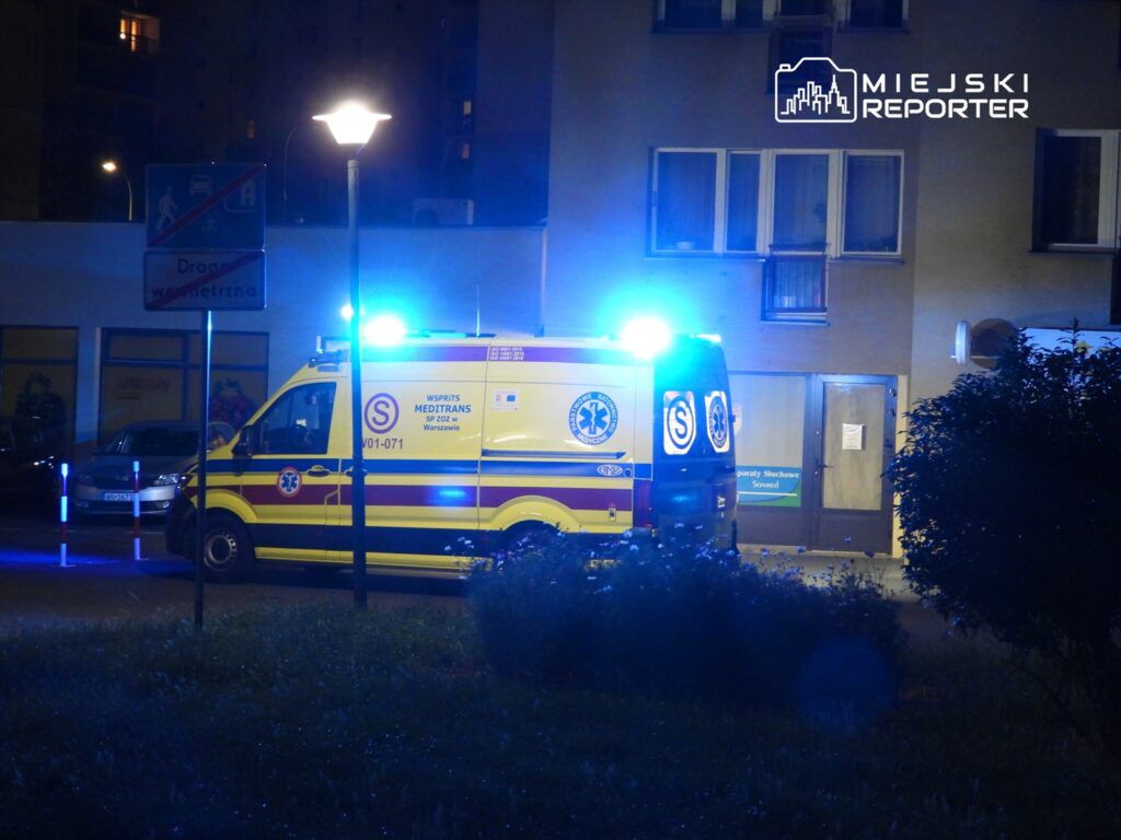 Karetka pogotowia z włączonymi sygnałami świetlnymi stoi na ulicy nocą, obok budynku mieszkalnego.
