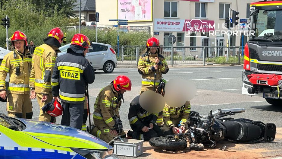 Na zdjęciu widać strażaków interweniujących na miejscu wypadku motocyklowego. Ratownicy udzielają pomocy poszkodowanemu, a obok leży przewrócony motocykl. W tle widoczny jest pojazd strażacki oraz ruch uliczny. Na miejscu zdarzenia widoczne są również oznaczenia służb ratunkowych.