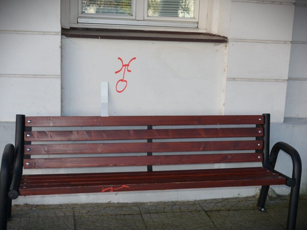Czerwony graffiti w kształcie znaku na białej ścianie obok drewnianej ławki w miejskim otoczeniu.