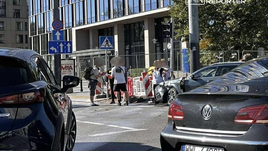 Grupa osób w białych koszulkach rozmawia obok uszkodzonego samochodu na ruchliwej ulicy w mieście.