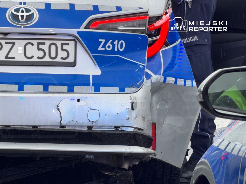 Policyjny radiowóz z uszkodzoną tylną częścią stoi na drodze, obok policjant w mundurze.