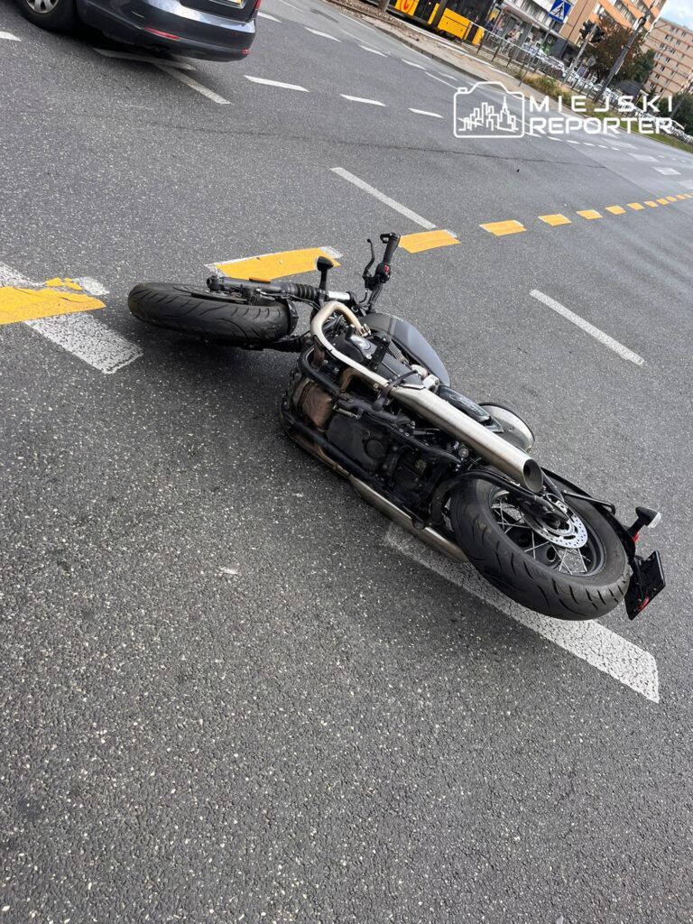 Motocykl leżący na boku na jezdni, w pobliżu skrzyżowania. W tle widoczny ruch uliczny, w tym samochody i tramwaj. Obraz ilustruje sytuację po wypadku drogowym, podkreślając zagrożenia związane z jazdą na dwóch kółkach. Brak osób w kadrze.