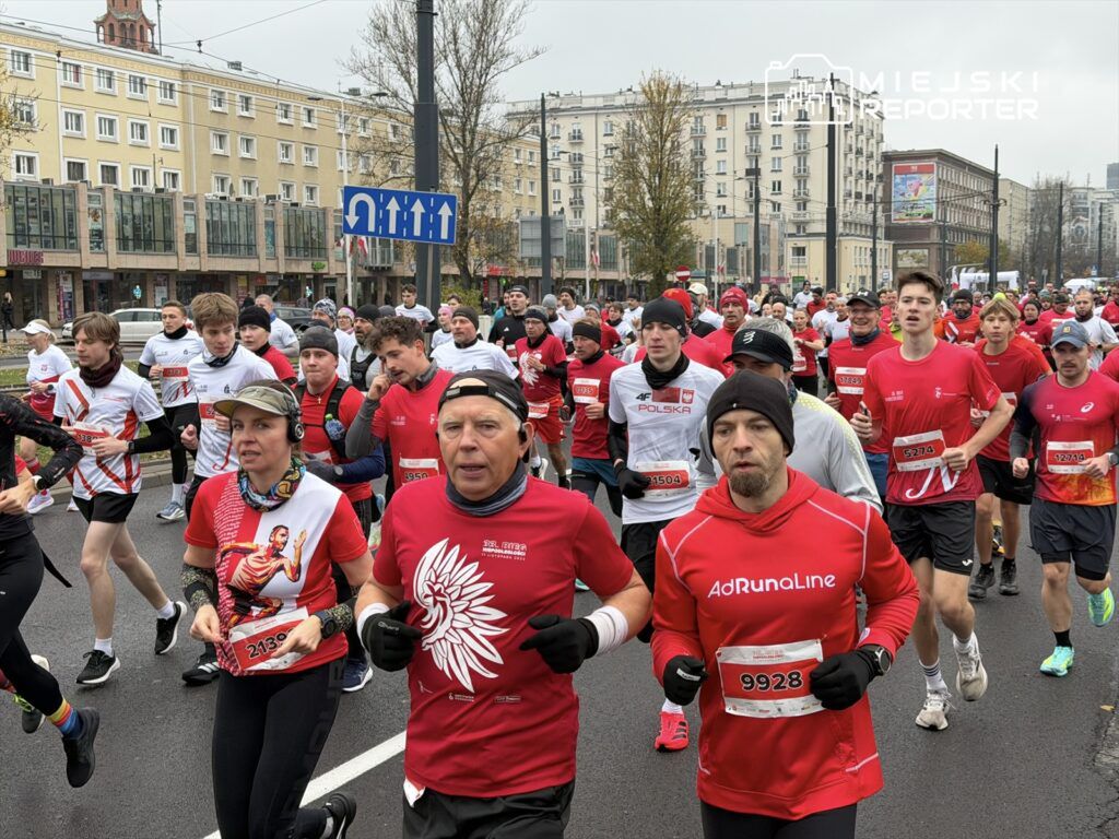 Biegacze w czerwonych koszulkach uczestniczą w wyścigu na miejskiej ulicy, obok nowoczesnych budynków.