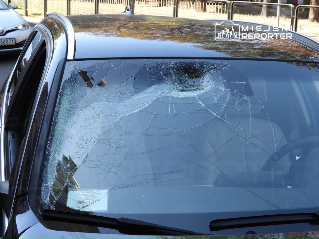 Pęknięta szyba przednia samochodu z licznymi rysami, znajdująca się na parkingu w miejskiej okolicy.