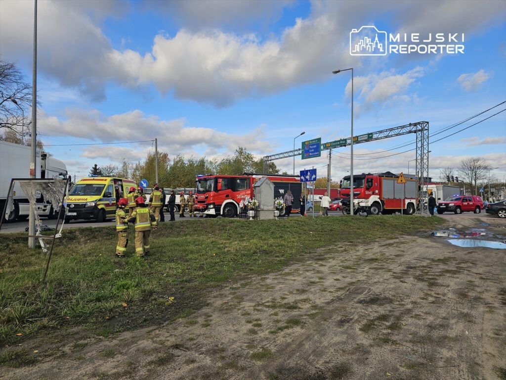 Grupa strażaków w mundurach idzie w stronę miejsca wypadku drogowego, gdzie stoją karetka i wozy strażackie.