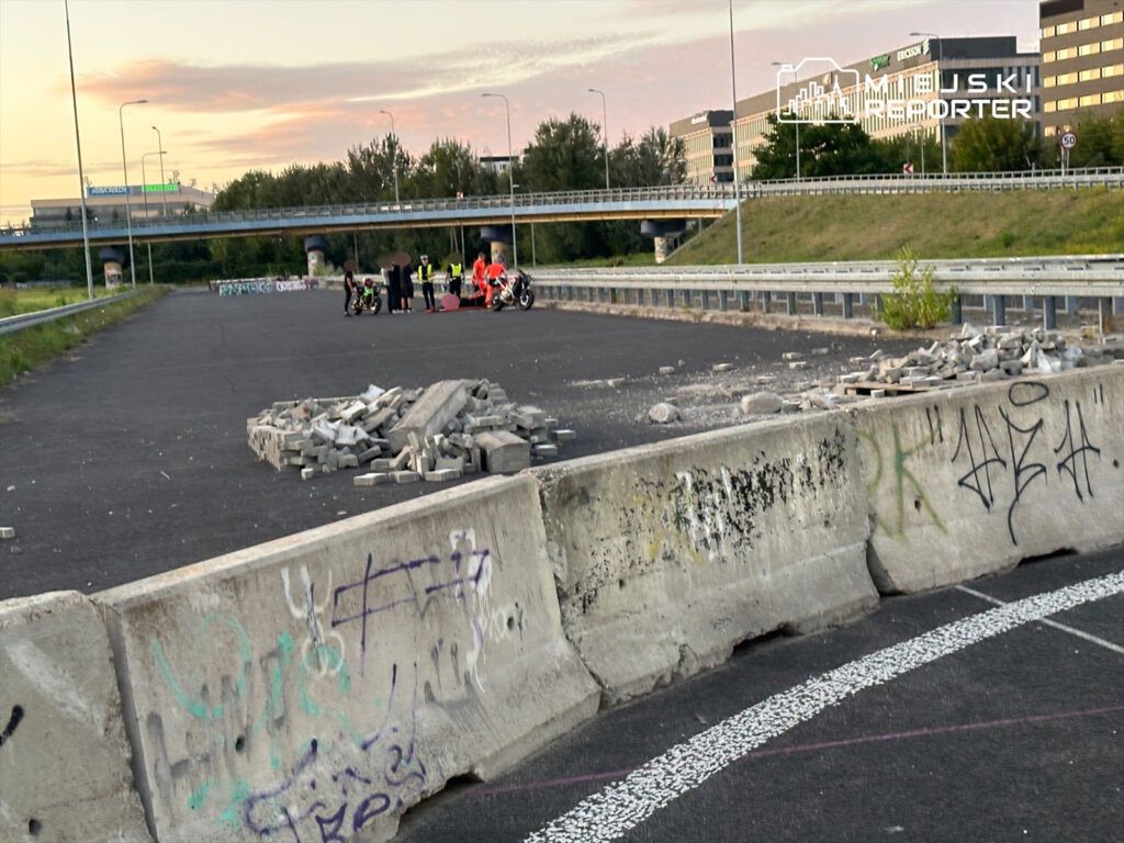 Grupa osób w kamizelkach odblaskowych zbiera się na pustym terenie z odłożonymi motocyklami, w tle widać graffiti i budynki.