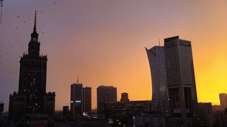 Zachód słońca nad miastem, z widokiem na wysokie budynki i charakterystyczną wieżę w tle.