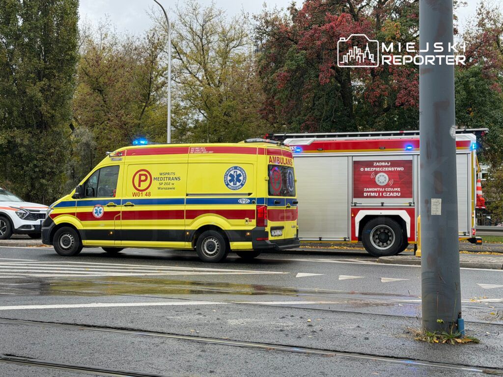 Karetka pogotowia z niebieskimi światłami zatrzymana obok wozu strażackiego na ulicy w deszczowy dzień.