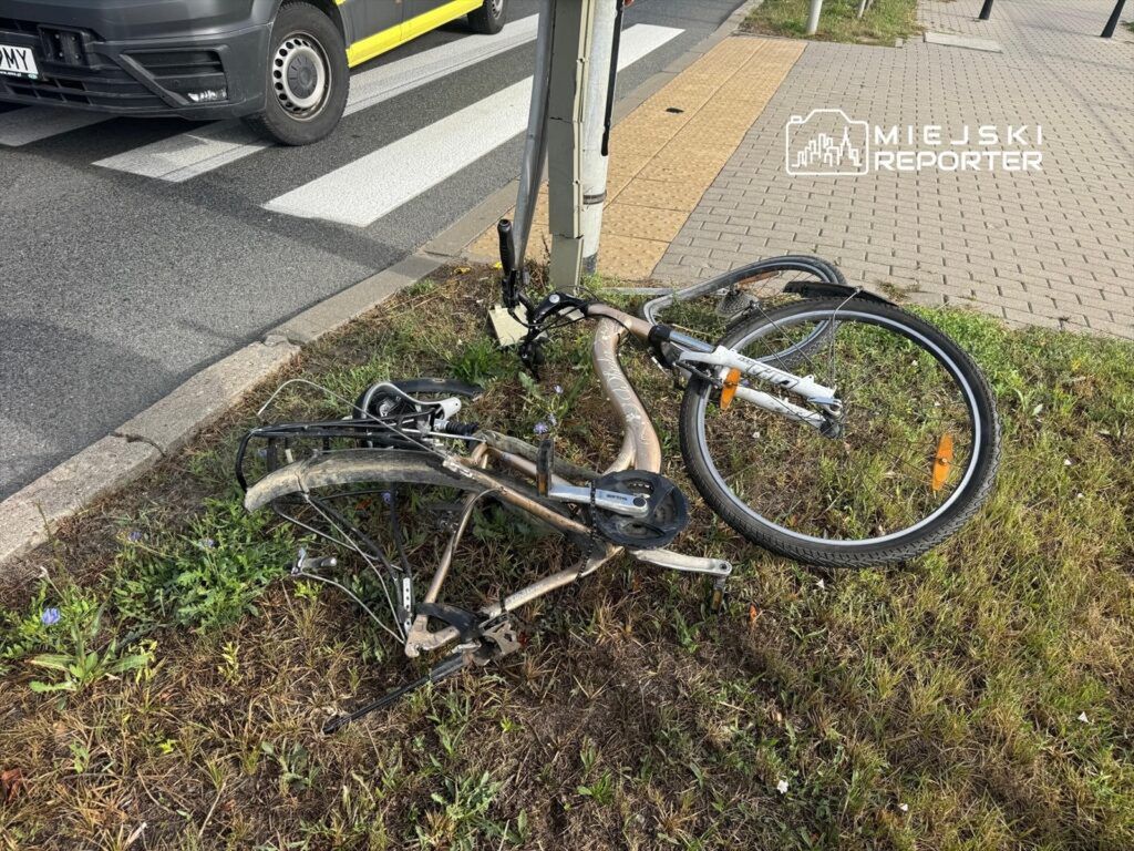 Złamany rower leży na trawie obok chodnika, obok przejścia dla pieszych i ruchliwej ulicy.