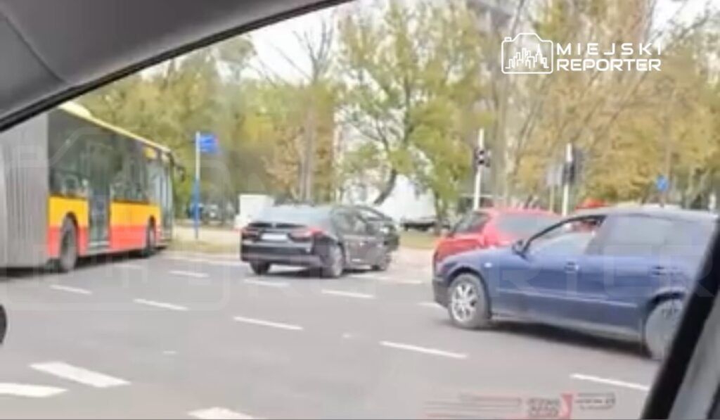 Ruchliwa ulica z samochodami, w tym autobusem, zatrzymującym się na przystanku. W tle drzewa i sygnalizacja świetlna.