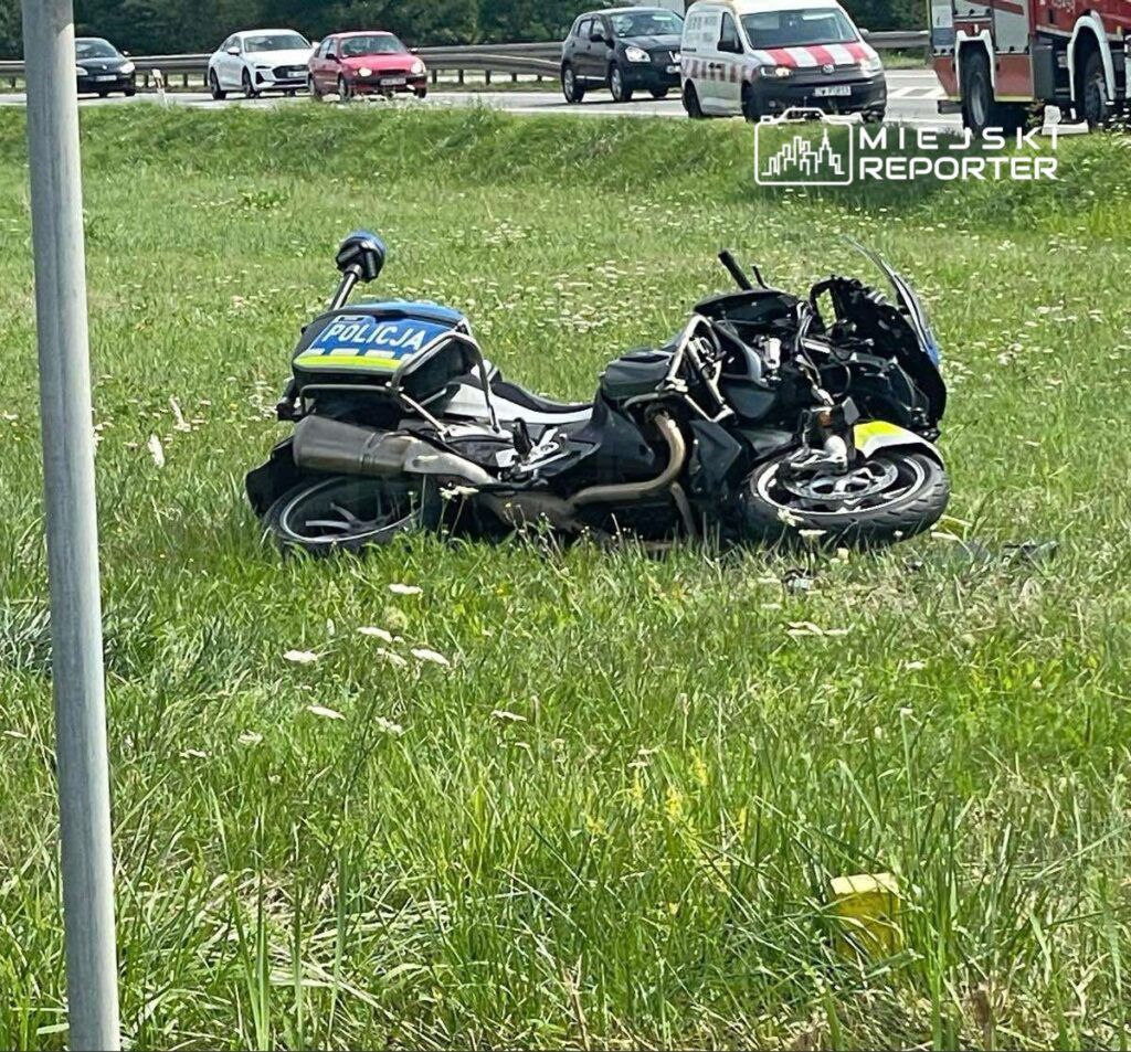 Zniszczony motocykl policyjny leży na trawie obok drogi, w tle widoczne pojazdy osobowe.