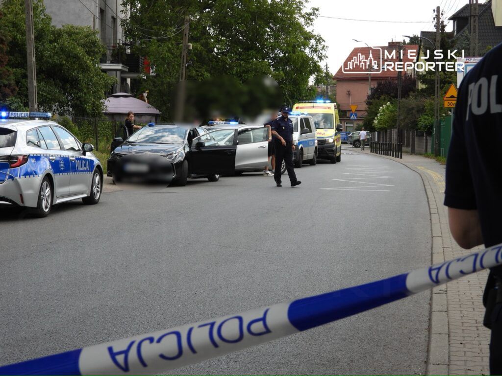 Grupa policjantów prowadzi interwencję na ulicy, obok zaparkowane są radiowozy oraz karetka pogotowia.