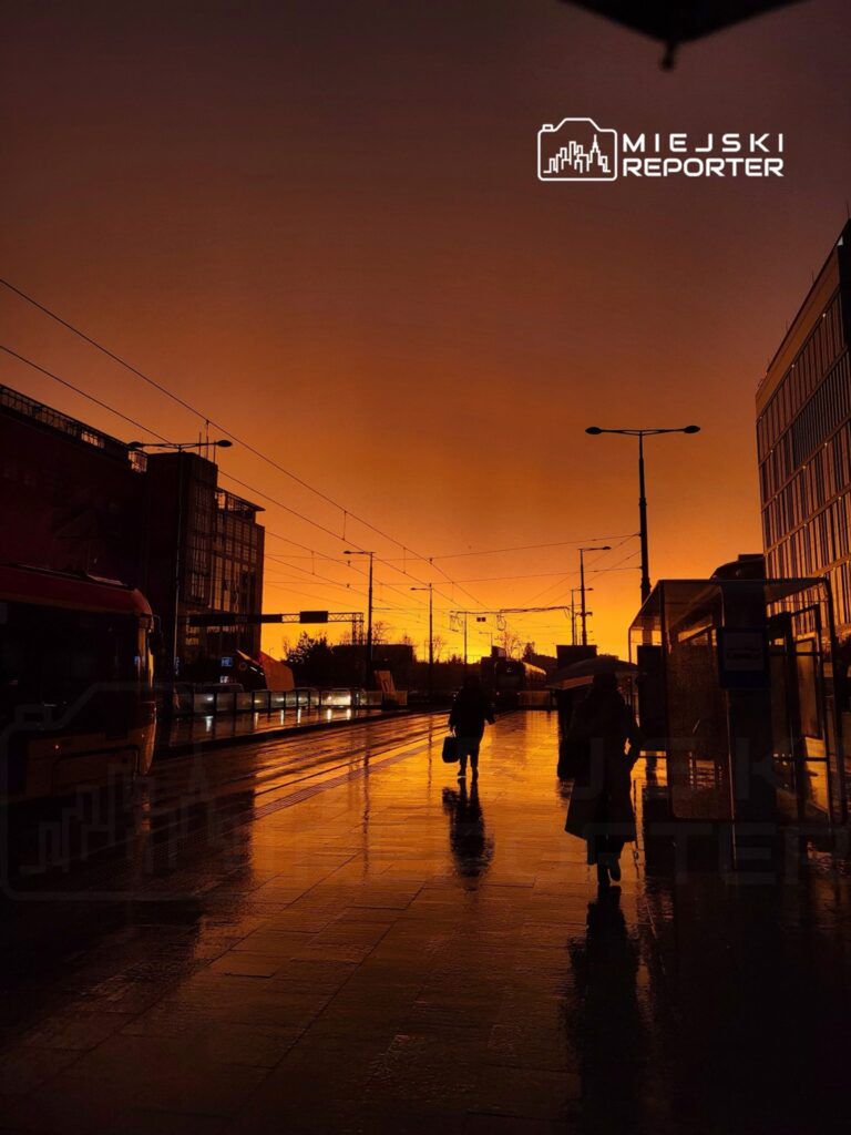 Osoby idące w deszczu po mokrej nawierzchni przystanku tramwajowego, w tle pomarańczowe niebo o zachodzie słońca.