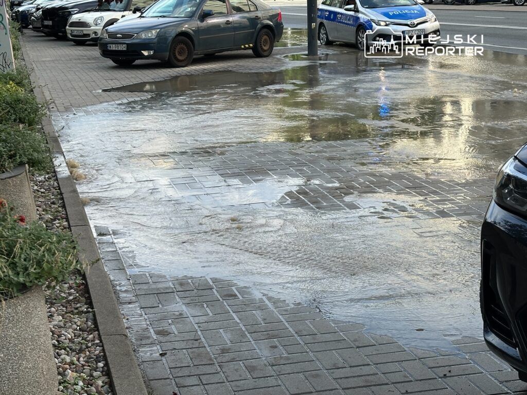 Woda rozlana na chodniku obok zaparkowanych samochodów, w tle radiowóz policyjny.