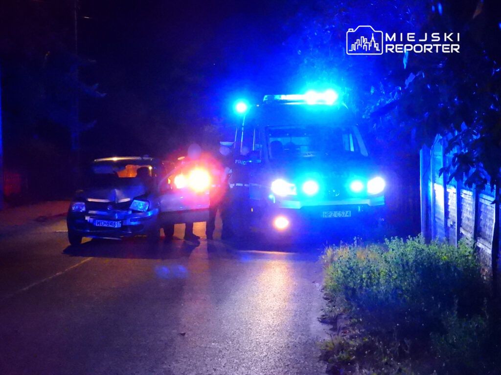 Policjanci rozmawiają z kierowcą przy uszkodzonym samochodzie na oświetlonej ulicy nocą.