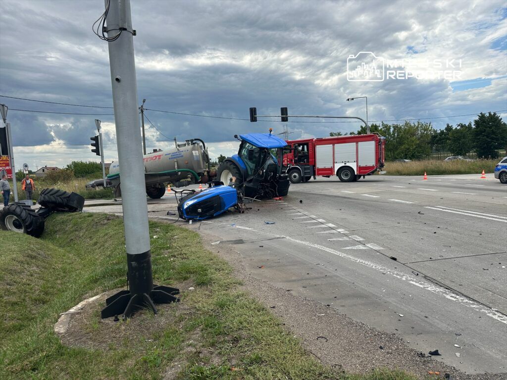 Traktor leży na boku obok drogi, a w tle widać wozy strażackie i osoby zabezpieczające miejsce wypadku.