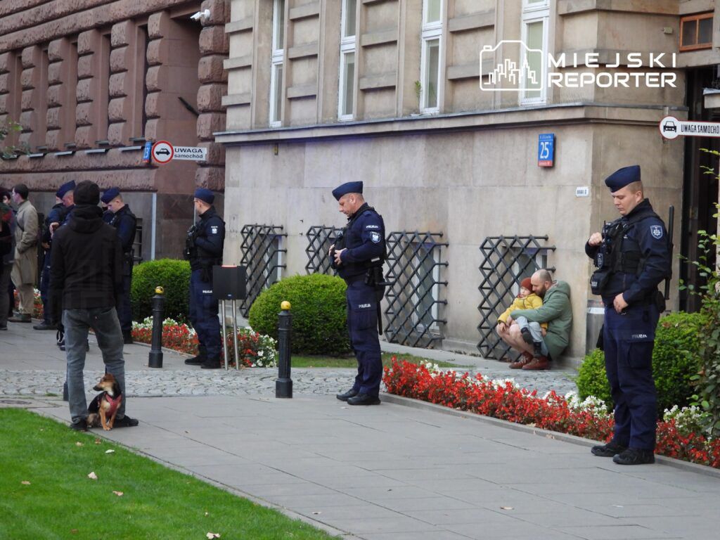 Grupa policjantów w mundurach stoi na chodniku obok budynku, a mężczyzna z dzieckiem siedzi na ziemi.