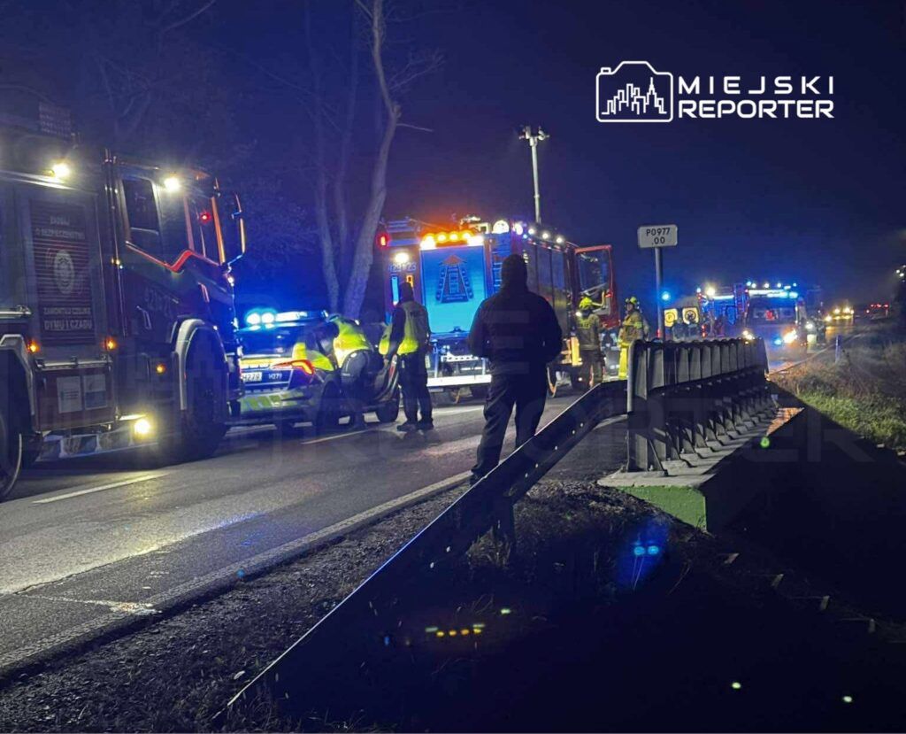 Ratownicy i policjanci pracują przy wypadku drogowym na ciemnej, oświetlonej ulicy, otoczeni przez pojazdy służb.